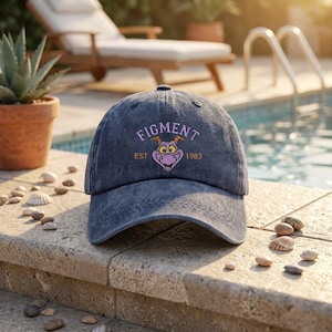 May include: A charcoal gray acid-wash baseball cap featuring an embroidered graphic of a purple dragon face with yellow horns. The text "FIGMENT" is arched above the dragon in light purple, with "EST 1983" in orange text on either side.