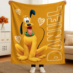 May include: Mustard yellow blanket featuring a cartoon Pluto character with a green collar and a long pink tongue. The blanket has white hearts and the name "DANIEL" in a stitched font. The blanket is held up by a person wearing white sneakers.