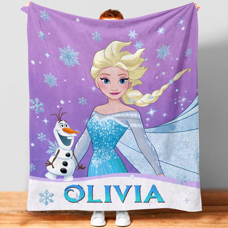 May include: A purple fleece blanket featuring a graphic print of a blonde character in a blue dress and a white snowman character. The background is decorated with light blue snowflakes and sparkles. The name OLIVIA is printed in blue letters.