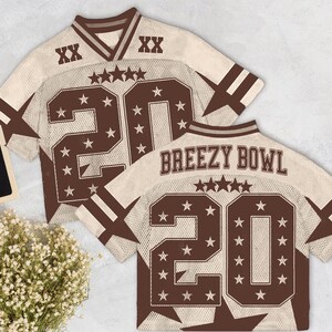 Pu&ograve; includere: Due maglie da football beige e marroni con il numero 20 e dettagli a stella. Una maglia ha la scritta "Breezy Bowl" sul retro. Le maglie hanno un aspetto vintage e usurato.