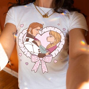 Puede incluir: Camiseta blanca con una ilustraci&oacute;n de dibujos animados de dos personajes dentro de un marco en forma de coraz&oacute;n. El dise&ntilde;o incluye un lazo rosa, corazones y los personajes mir&aacute;ndose. La camiseta es de material suave.