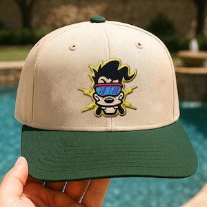 May include: A tan baseball cap with a green brim and a green button on top. The cap features an embroidered cartoon character with black hair, blue goggles, and yellow lightning bolts.
