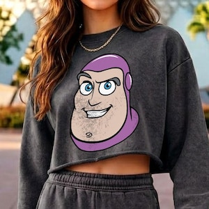 May include: A charcoal grey mineral-wash cropped sweatshirt featuring a large graphic print of a cartoon character with a purple hood, blue eyes, and a prominent chin. The long-sleeved garment has a raw-cut hem and a crew neckline.