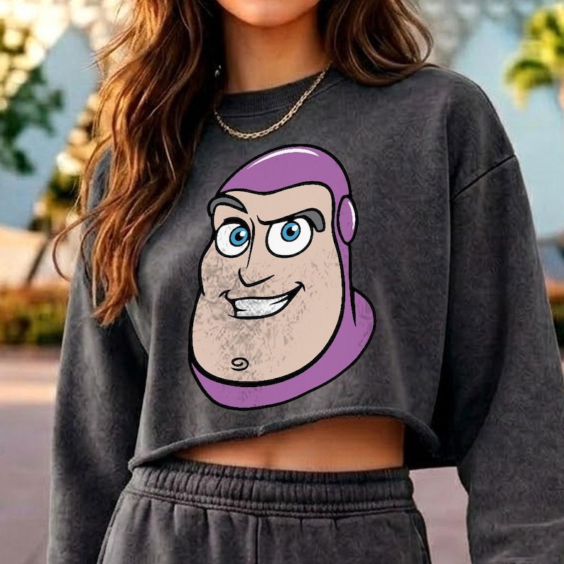 May include: A charcoal grey mineral-wash cropped sweatshirt featuring a large graphic print of a cartoon character with a purple hood, blue eyes, and a prominent chin. The long-sleeved garment has a raw-cut hem and a crew neckline.