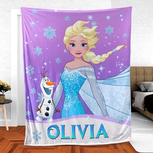 May include: A purple fleece blanket featuring a graphic print of Elsa with blonde hair and a blue dress, alongside Olaf the snowman. The background is decorated with light blue snowflakes and sparkles. The name OLIVIA is printed in blue letters.