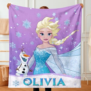 May include: A purple fleece blanket featuring a graphic print of Elsa with blonde hair and a blue dress, alongside Olaf the snowman. The background is decorated with light blue snowflakes and sparkles. The name OLIVIA is printed in blue letters.