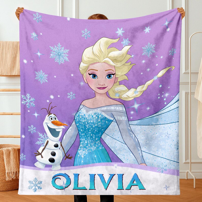 May include: A purple fleece blanket featuring a graphic print of Elsa with blonde hair and a blue dress, alongside Olaf the snowman. The background is decorated with light blue snowflakes and sparkles. The name OLIVIA is printed in blue letters.