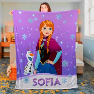 May include: A purple fleece blanket featuring a printed illustration of Anna from Frozen and Olaf the snowman. The design includes light blue snowflakes and stars scattered across the fabric. The name SOFIA is printed in large purple letters.