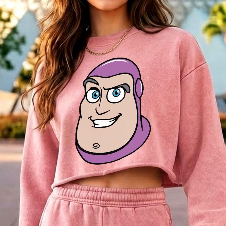 May include: A dusty pink long-sleeve cropped sweatshirt featuring a large, centred graphic print of the character Buzz Lightyear. The sweatshirt has a crew neckline, long sleeves, and a raw-cut hemline, paired with matching pink sweatpants.