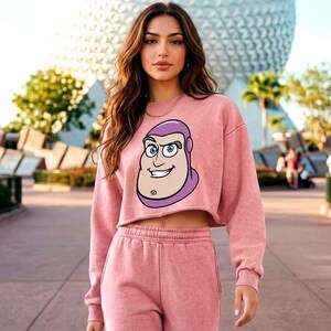 May include: A dusty rose cropped sweatshirt featuring a graphic print of the character Buzz Lightyear on the front, paired with matching high-waisted sweatpants. The sweatshirt has a crew neckline, long sleeves, and a raw-edge hemline.