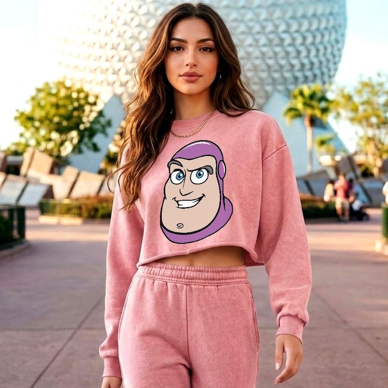 May include: A dusty rose cropped sweatshirt featuring a graphic print of the character Buzz Lightyear on the front, paired with matching high-waisted sweatpants. The sweatshirt has a crew neckline, long sleeves, and a raw-edge hemline.