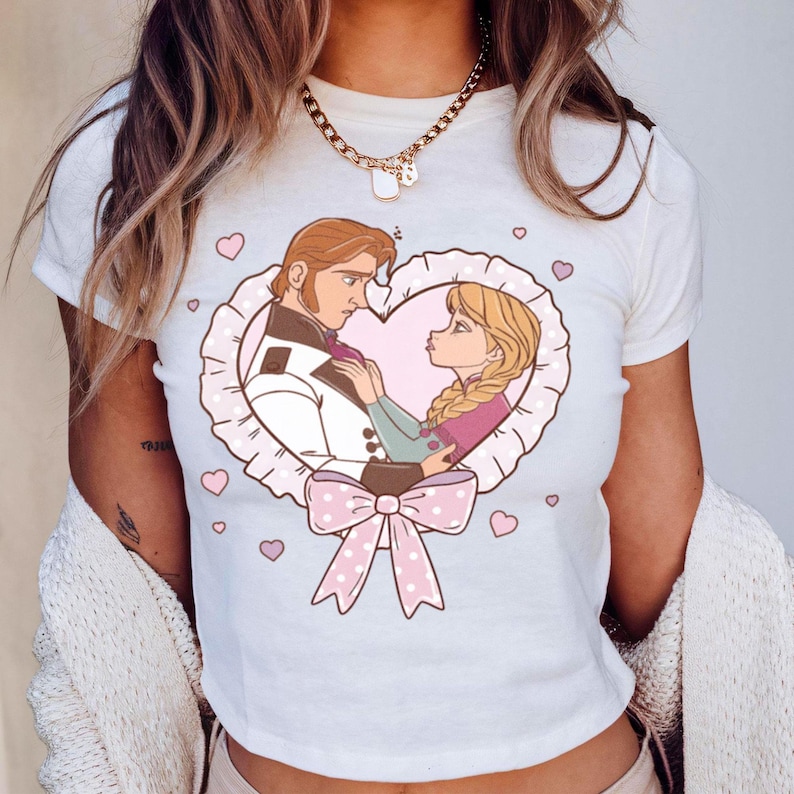 Puede incluir: Camiseta blanca con una ilustraci&oacute;n de dibujos animados de una pareja en un marco en forma de coraz&oacute;n con un lazo rosa. La ilustraci&oacute;n representa a un hombre con traje blanco y una mujer con trenzas rubias. Corazones rosas rodean el marco.