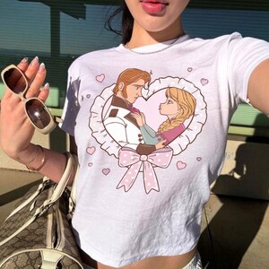 Puede incluir: Top corto blanco con una ilustraci&oacute;n de dibujos animados de una pareja dentro de un marco en forma de coraz&oacute;n con un lazo rosa. El dise&ntilde;o incluye corazones rosas. Prenda casual para uso diario.