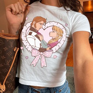 Puede incluir: Camiseta blanca con una ilustraci&oacute;n de dibujos animados de una pareja dentro de un marco en forma de coraz&oacute;n. El dise&ntilde;o incluye un lazo rosa, corazones y la pareja en una pose rom&aacute;ntica. La camiseta es de material suave y es adecuada para uso casual.