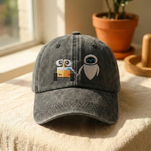 May include: A charcoal grey washed denim baseball cap featuring an embroidered graphic of two robots, one yellow and one white, holding hands. The cap has a curved brim, a six-panel construction with stitched eyelets, and a structured front crown.