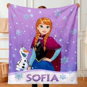 May include: A purple fleece blanket featuring a graphic print of the character Anna with braided hair and the snowman Olaf. The background is decorated with light blue snowflakes and sparkles. The name SOFIA is printed in large purple letters.