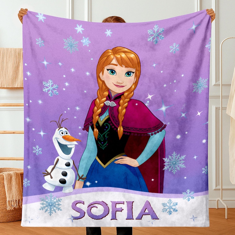 May include: A purple fleece blanket featuring a graphic print of the character Anna with braided hair and the snowman Olaf. The background is decorated with light blue snowflakes and sparkles. The name SOFIA is printed in large purple letters.
