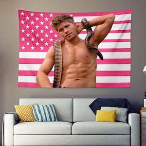 May include: A pink and white American flag tapestry with a shirtless person and a snake draped around their neck. The tapestry is hanging on a wall above a white sofa with decorative pillows.