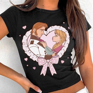 Puede incluir: Top corto negro con una ilustraci&oacute;n de dibujos animados de una pareja dentro de un marco en forma de coraz&oacute;n. El coraz&oacute;n est&aacute; rodeado por un borde blanco similar al encaje y un lazo rosa. Peque&ntilde;os corazones rosas est&aacute;n dispersos alrededor del dise&ntilde;o principal.