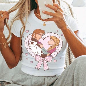 Puede incluir: Camiseta blanca con una ilustraci&oacute;n de dibujos animados de una pareja dentro de un marco en forma de coraz&oacute;n, adornada con un lazo rosa. La imagen muestra a un hombre con traje blanco y una mujer con trenzas rubias. Corazones rosas dispersos.