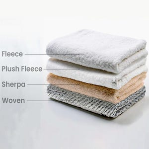 May include: A stack of four folded fabric swatches labelled with text. The top layer is white fleece, the second is white plush fleece, the third is tan sherpa, and the bottom layer is a grey and white woven fabric with a visible grid-like texture.