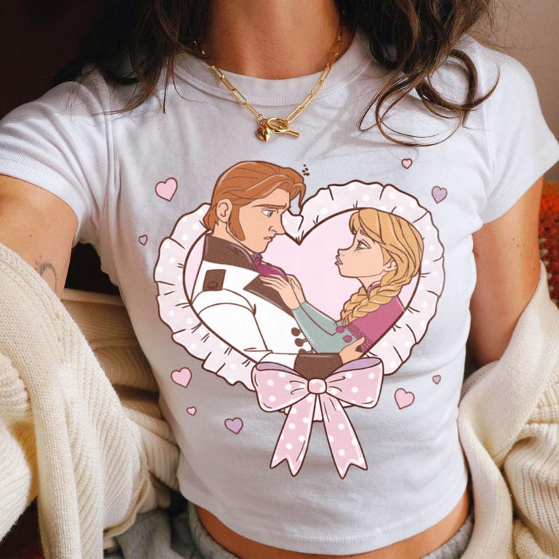 Puede incluir: Camiseta blanca con una ilustraci&oacute;n de dibujos animados de una pareja en un marco en forma de coraz&oacute;n. La imagen muestra a un hombre y una mujer en una pose rom&aacute;ntica, rodeados de corazones rosas y un lazo de lunares.