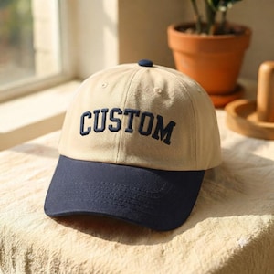 May include: A two-tone baseball cap featuring a tan crown and a dark navy blue brim. The word CUSTOM is embroidered in dark navy blue block lettering across the front panels of the cap. The hat includes a small dark navy blue button on top.