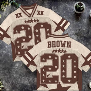 Pu&ograve; includere: Due maglie da football marroni e color crema. Ogni maglia ha il numero 20 in grandi lettere marroni con dettagli a stella. La parola "BROWN" &egrave; stampata sul retro di una maglia. Le maglie hanno strisce marroni sulle maniche.
