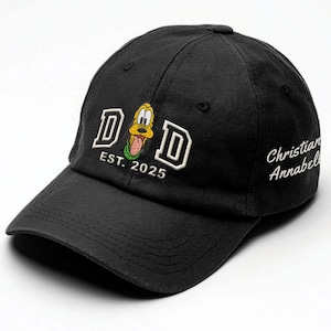 May include: Black baseball cap with a white embroidered "DAD" with a cartoon dog in the middle. The text "EST. 2025" is below the dog. The text "Christian Annabelle" is embroidered on the right side of the cap.