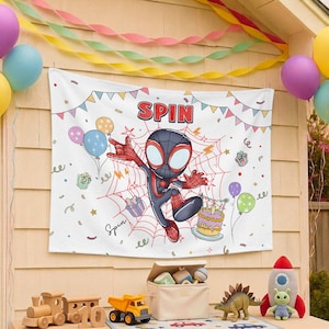 May include: A white rectangular fabric banner featuring a cartoon character in a black and red suit centered on a red spiderweb. The text SPIN appears at the top, surrounded by colorful illustrations of balloons, gift boxes, and a birthday cake.