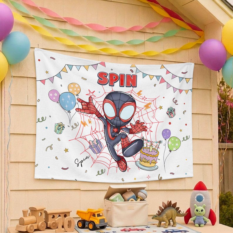May include: A white rectangular fabric banner featuring a cartoon character in a black and red suit centered on a red spiderweb. The text SPIN appears at the top, surrounded by colorful illustrations of balloons, gift boxes, and a birthday cake.