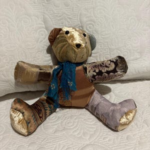 Puede incluir: Un oso de peluche de patchwork hecho a mano con una bufanda azul. El cuerpo del oso está hecho de varias telas estampadas en tonos dorados, marrones y morados. El oso está sobre una superficie acolchada blanca.
