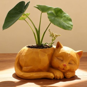 May include: A yellow cat-shaped planter with a sleeping cat design. The planter holds a green plant with large leaves. The cat has closed eyes and a pink nose. The planter is made of a textured material and sits on a wooden surface.