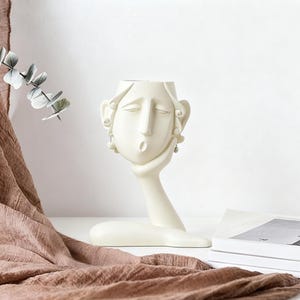 Puede incluir: Un jarrón decorativo de color crema con forma de rostro humano estilizado, con los ojos cerrados y una mano que sostiene la barbilla. El jarrón tiene un acabado liso y presenta detalles de pendientes. El diseño es moderno y artístico, adecuado para la decoración del hogar.