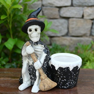 Op de afbeelding: Een Halloween-thema keramische skeletfiguur met een zwarte heksenhoed met een oranje band en een zwarte cape met witte spikkels. Het skelet houdt een bezem vast en zit naast een zwart-witte gespikkelde plantenbak.