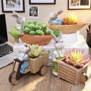 May include: A collection of decorative bunny figurines with planters. The bunnies are in various poses, some pushing wheelbarrows, others riding scooters, and one relaxing in a boat-shaped planter. The planters hold succulents and faux flowers.