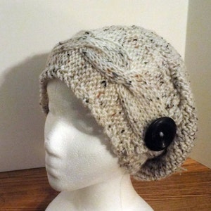 May include: A white knitted beanie with a cable knit pattern and a black button. The beanie is worn on a white mannequin head.