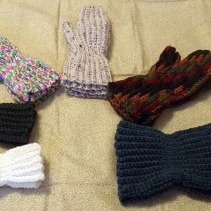 May include: A collection of six handmade crochet mittens in various colors. The mittens include a pair in black, white, and navy blue. Other pairs feature colorful patterns with pink, green, and yellow. The mittens are arranged on a light brown surface.