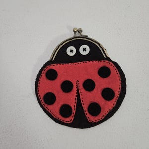 May include: A ladybird-shaped coin purse made of black felt with a red felt overlay. The red overlay features black circular spots. The purse has a metal clasp and button eyes.