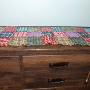 May include: A patchwork rag quilt in a rectangular shape, featuring squares of various plaid fabrics in red, green, purple, and tan. The quilt is placed on a dark wooden dresser.