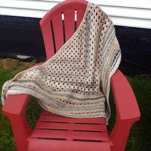 May include: A crocheted shawl in shades of beige, brown, and gray draped over a red plastic chair. The shawl has a square pattern and a fringed edge.