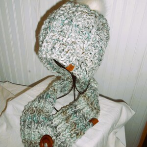 May include: A white and green knitted hat with a white fur pom pom and a matching knitted scarf with a wooden button.