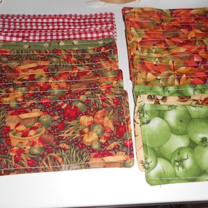 May include: A collection of fabric placemats with various patterns. Designs include red and white gingham, autumn leaves, green apples, and a harvest scene with fruits and vegetables. The placemats are neatly stacked.