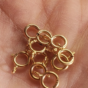 May include: Close-up of several small, gold-colored jewelry clasps. The clasps are circular with a spring-loaded mechanism for secure closure. The clasps are likely used for necklaces, bracelets, or other jewelry items.