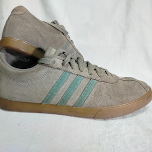 Può includere: Un paio di scarpe Adidas in pelle scamosciata grigia con strisce verdi. Le scarpe hanno una suola in gomma marrone.