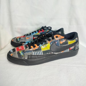 May include: Black and white Nike sneakers with a patchwork design featuring the Nike logo and the word "Nike" repeated throughout. The sneakers have black laces and a black sole.