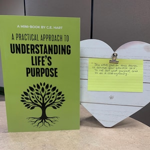 May include: A green mini-book titled "A Practical Approach to Understanding Life's Purpose" with a tree graphic. Beside it is a white heart-shaped wooden decoration with a yellow sticky note that reads "You were put on this earth to achieve your greatest self."