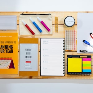 May include: A wooden board displaying stationery and planning tools. Includes a book titled "A Practical Approach to Planning Your Year", a notebook, sticky notes, markers, and a small whiteboard. The items are arranged for organization.
