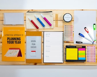 May include: A wooden board displaying stationery and planning tools. Includes a book titled "A Practical Approach to Planning Your Year", a notebook, sticky notes, markers, and a small whiteboard. The items are arranged for organization.
