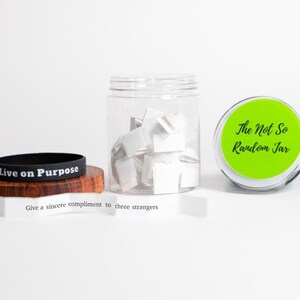 May include: An image featuring a black wristband with "Live on Purpose" printed on it, a clear jar filled with white slips of paper, and a jar with a green lid labeled "The Not So Random Jar". A slip of paper reads "Give a sincere compliment to three strangers".